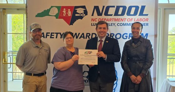 Four people pose for a photo at a Safety Award program, with a certificate being presented and an NCDOL banner in the background.